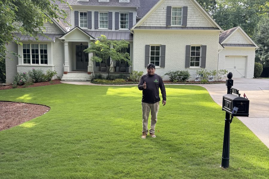 A crew member stands in front of a large house with a newly installed, vibrant green lawn